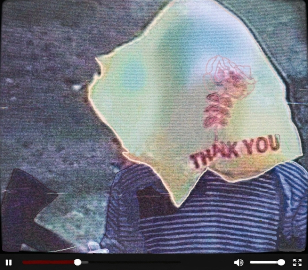 A photo of an unknown person standing in front of the camera, holding an axe. There is a yellow plastic bag over their head with a red text and a rose that reads "THANK YOU". There is VHS grain on the photo.